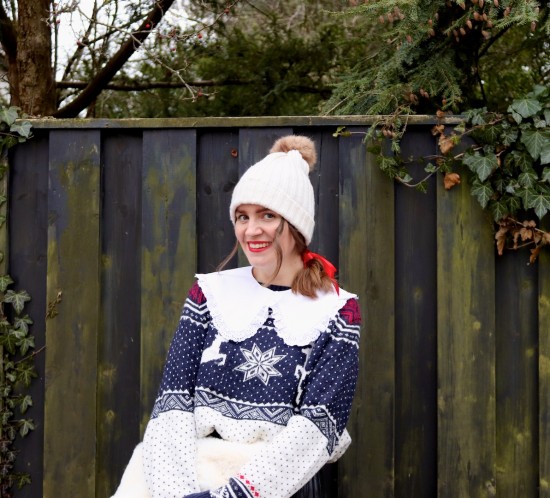 Cozy Chic Holiday Look. Fashion Blogger Girl by Style Blog Heartfelt Hunt. Girl with blond hair and a red bow wearing a pompom beanie, white collar, cozy sweater, pleated skirt, faux fur bag and combat boots.