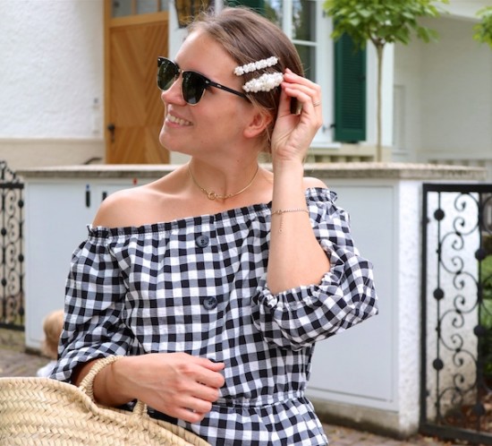 Gingham Off Shoulder Top. Fashion Blogger Girl by Style Blog Heartfelt Hunt. Girl with blond low messy bun and pearl hair clips wearing a gingham off shoulder top, Ray-Ban sunglasses, floral ruffled skirt, straw bag and black sandals.