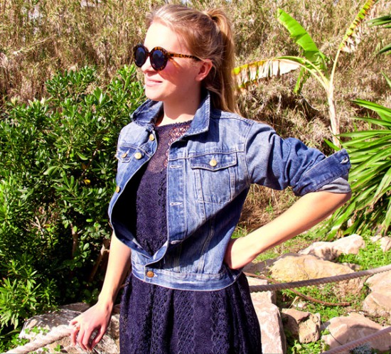 Smiling blond fashion and style blogger girl with high ponytail wearing a dark-blue lace dress, denim jacket and sunglasses