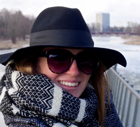 Smiling blond fashion and style blogger girl in closeup view wearing an oversized jacket, black hat, sunglasses and a black and white scarf