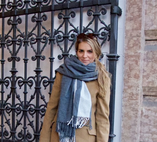 Blonde fashion blogger girl with sunglasses, oversized scarf and camel coat standing in front of a gate