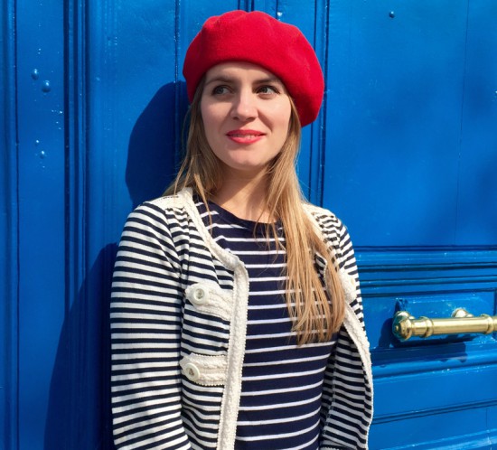 Smiling blonde fashion and style blogger girl in closeup view wearing different stripes, red béret and red lipstick in front of a blue door