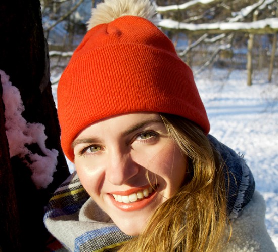 Smiling blonde fashion and style blogger girl with orange beanie, plaid scarf and oversized winter coat in a beautiful, snowy scenery