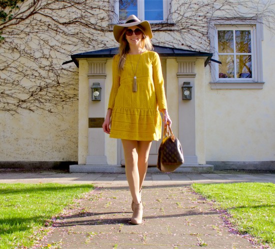 Smiling blonde fashion and style blogger girl wearing a yellow lace dress, sunglasses, floppy hat, fringe necklace, boots and a classic Louis Vuitton Monogram bag