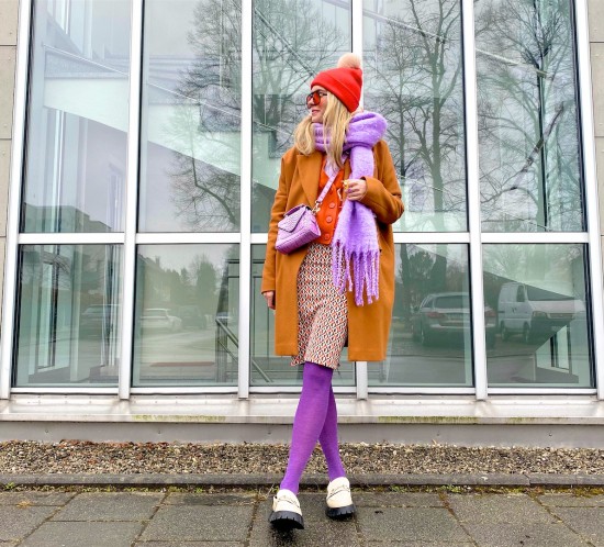 Lilac Orange. Fashion Blogger Girl by Style Blog Heartfelt Hunt. Girl with blond hair wearing a lilac scarf, orange sunglasses, orange beanie, orange coat, orange cardigan, orange jacquard skirt, purple tights and chunky loafers.