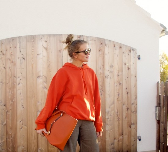 Orange Hoodie. Fashion Blogger Girl by Style Blog Heartfelt Hunt. Girl with blond high, messy bun wearing an orange hoodie, flared pants, Ray-Ban sunglasses, orange Michael Kors bag and heeled boots.