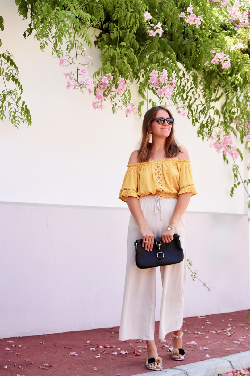 Pompon Straw Sandals. Fashion Blogger Girl by Style Blog Heartfelt Hunt. Girl with blond hair wearing a yellow off shoulder top, wide pants, tortoise shell sunglasses, tassel earrings, clutch and pompon straw sandals.