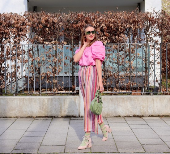 Rainbow Pants. Fashion Blogger Girl by Style Blog Heartfelt Hunt. Girl with blond hair wearing rainbow pants, pink puff sleeve blouse, green sunglasses, green ruched bag, green socks and pink heels.
