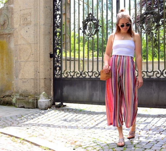 Rainbow Pants. Fashion Blogger Girl by Style Blog Heartfelt Hunt. Girl with blond hair wearing a rainbow pants, white top, Ray-Ban sunglasses, straw bag and glittery sandals.