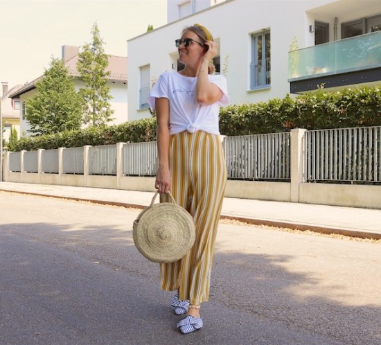 Yellow Stripes. Fashion Blogger Girl by Style Blog Heartfelt Hunt. Girl with blond, low messy bun and yellow headband wearing a jumpsuit with yellow stripes, white T-shirt, Ray-Ban sunglasses, round straw bag and striped mules.